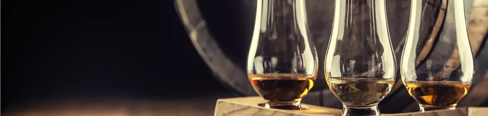 Drei Whiskygläser mit unterschiedlichen Bernsteintönen auf einem Holzständer vor einem dunklen Hintergrund mit einem Fass im Hintergrund.